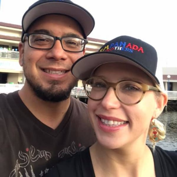 A man and woman smiling, both wearing Empanada Brothers hats, with water in the background.