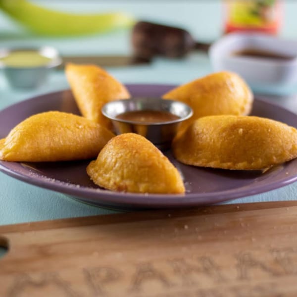 Five golden Colombian empanadas arranged on a purple plate with a small bowl of dipping sauce.