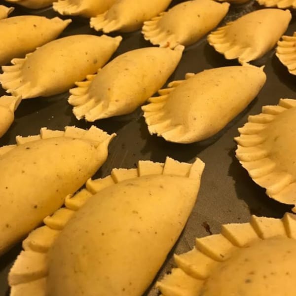 Rows of uncooked Colombian empanadas with crimped edges on a dark baking sheet, ready for frying.