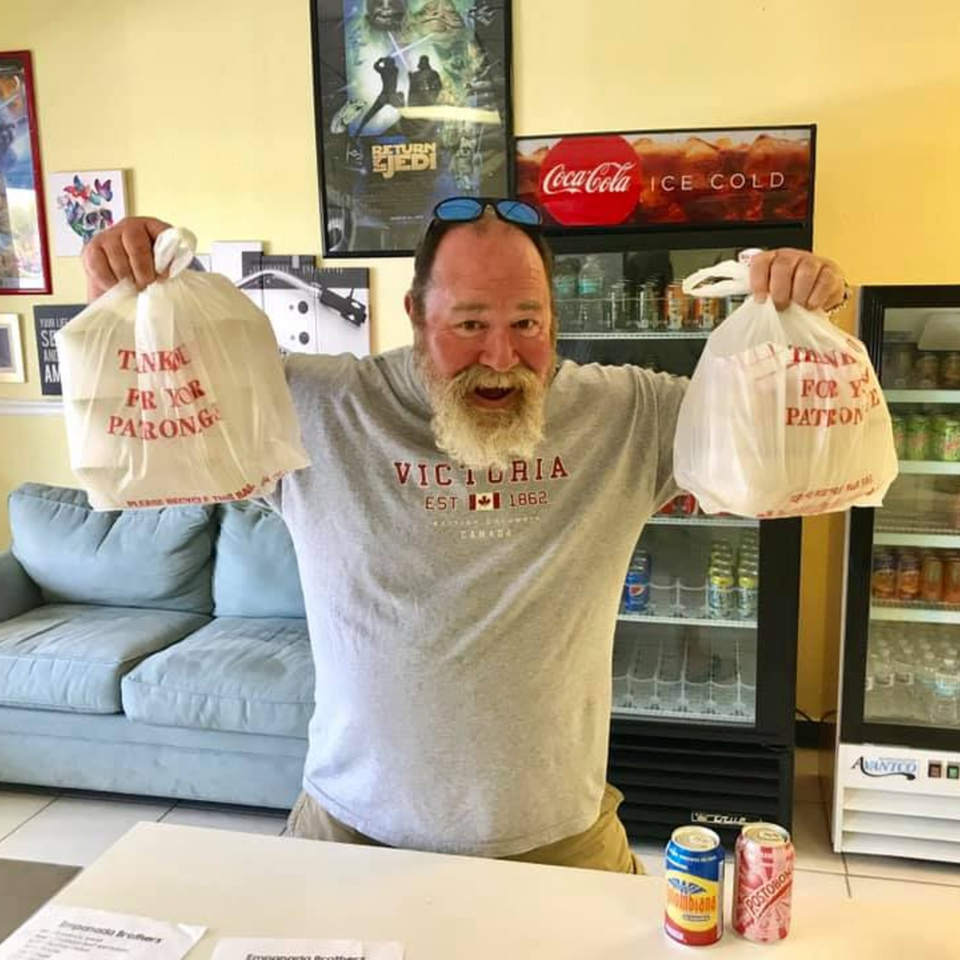 A smiling man holds two Empanada Brothers takeout bags, with Colombian sodas on the counter.