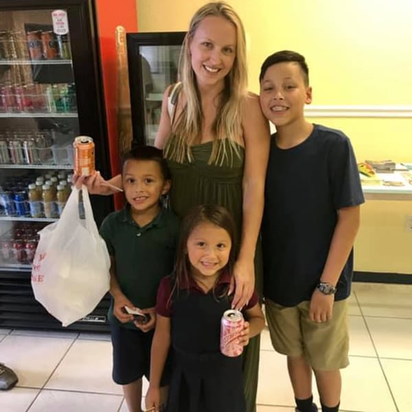 A woman and three children smiling, holding drinks and a bag, in front of a refrigerated display.