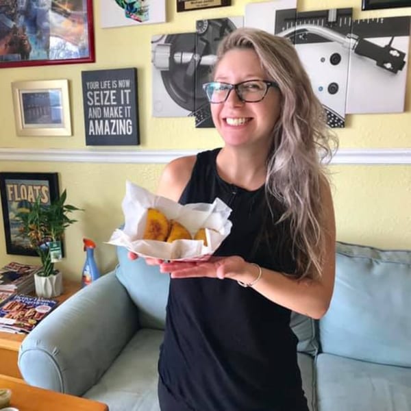 Smiling woman in glasses holding a basket of Empanada Brothers' Colombian empanadas in a living room.