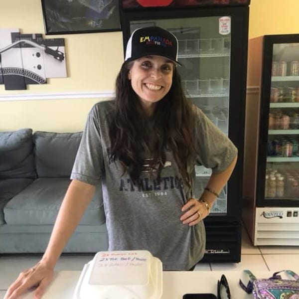 Smiling woman in an Empanada Brothers hat stands behind a counter with refrigerators and a couch.