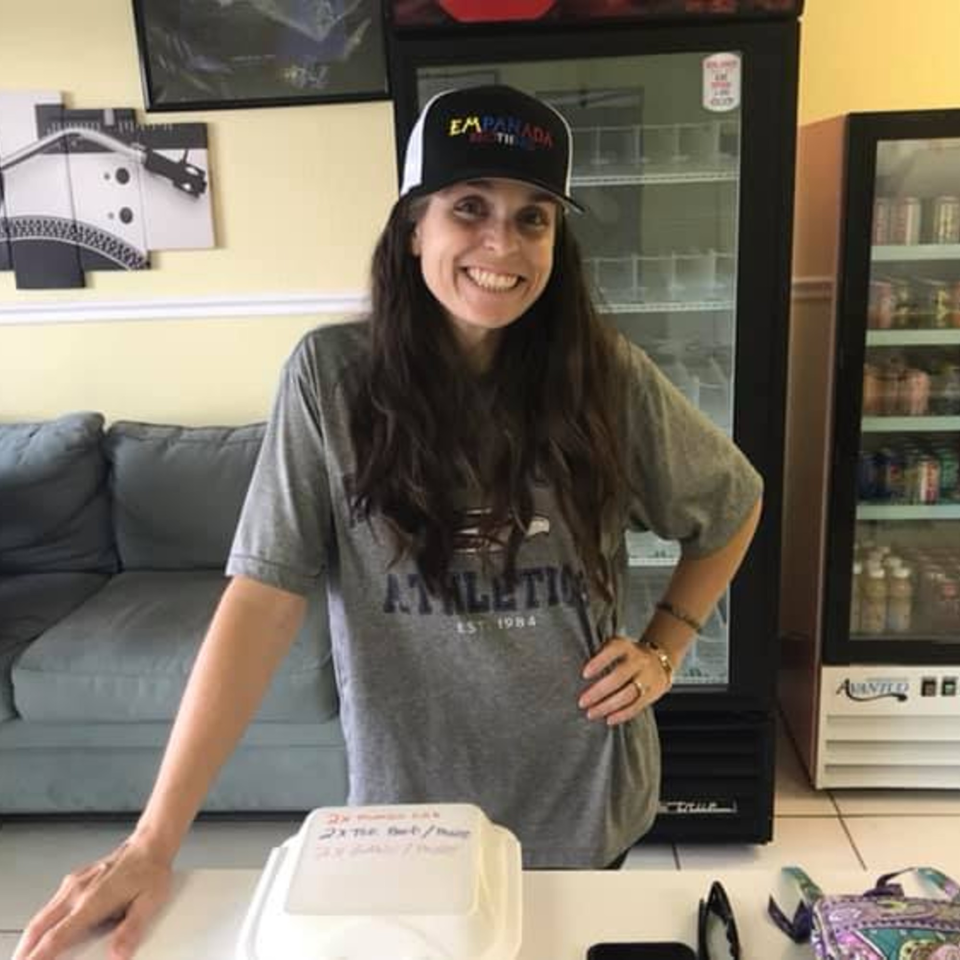 Smiling woman in an Empanada Brothers hat stands behind a counter with refrigerators and a couch.
