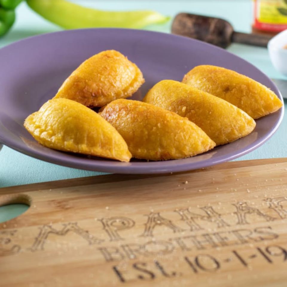 Six golden-brown Empanada Brothers empanadas on a purple plate, with a branded wooden board below.