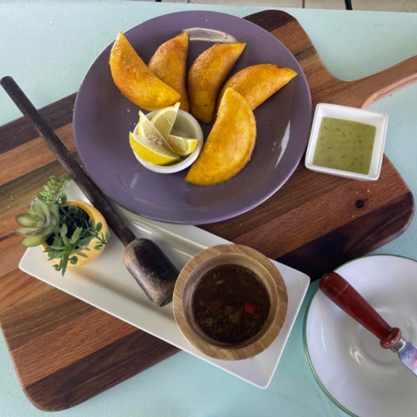 Five golden Empanada Brothers empanadas with lemon wedges, green sauce, and dark sauce on a wooden board.