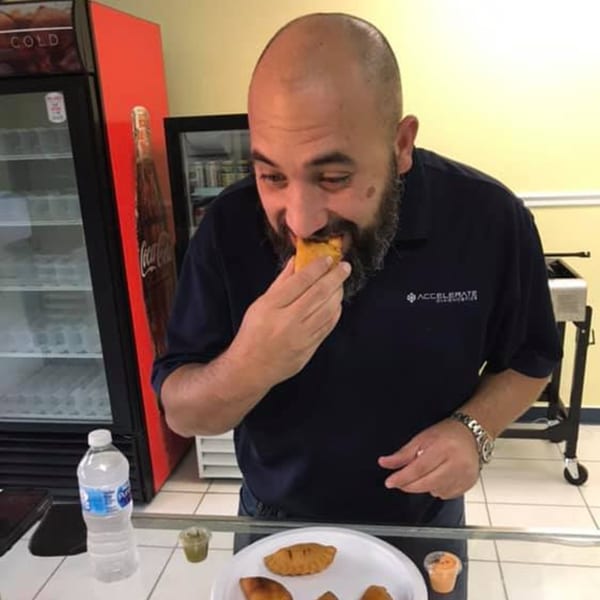A man in a dark polo shirt takes a bite of a golden-brown Empanada Brothers empanada.