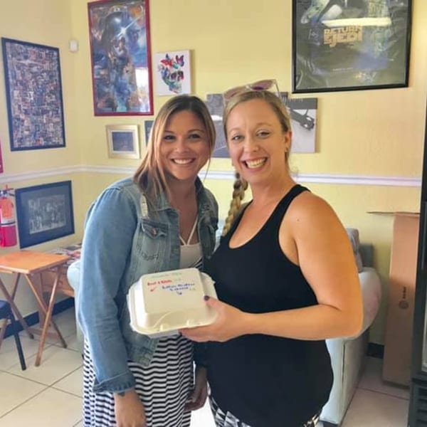 Two smiling women, one in a denim jacket and striped dress, holding a takeout container.