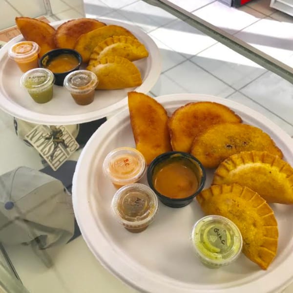 Two plates of golden-fried Colombian empanadas with various dipping sauces on a white tiled surface.